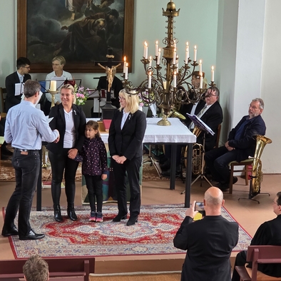 Zwei Frauen stehen vor einem Altar in einer Kirche, umgeben von Mitgliedern des Posaunenchores und ihnen wird gratuliert von einem Mann.