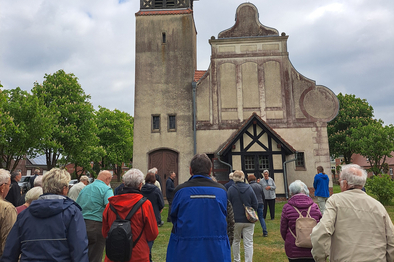 Start der Kirchbaufahrt in Helle Viele Besucher stehen auf dem Rasen vor der Kirche in Helle und hören einer Frau zu, die etwas erklärt.