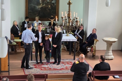 Zwei Frauen stehen vor einem Altar in einer Kirche, umgeben von Mitgliedern des Posaunenchores und ihnen wird gratuliert von einem Mann.