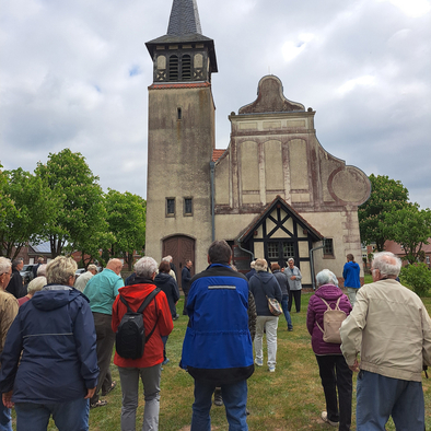 Viele Besucher stehen auf dem Rasen vor der Kirche in Helle und hören einer Frau zu, die etwas erklärt.
